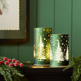 Two green tree luminaries glowing on a dark mantel with pine and red berries
