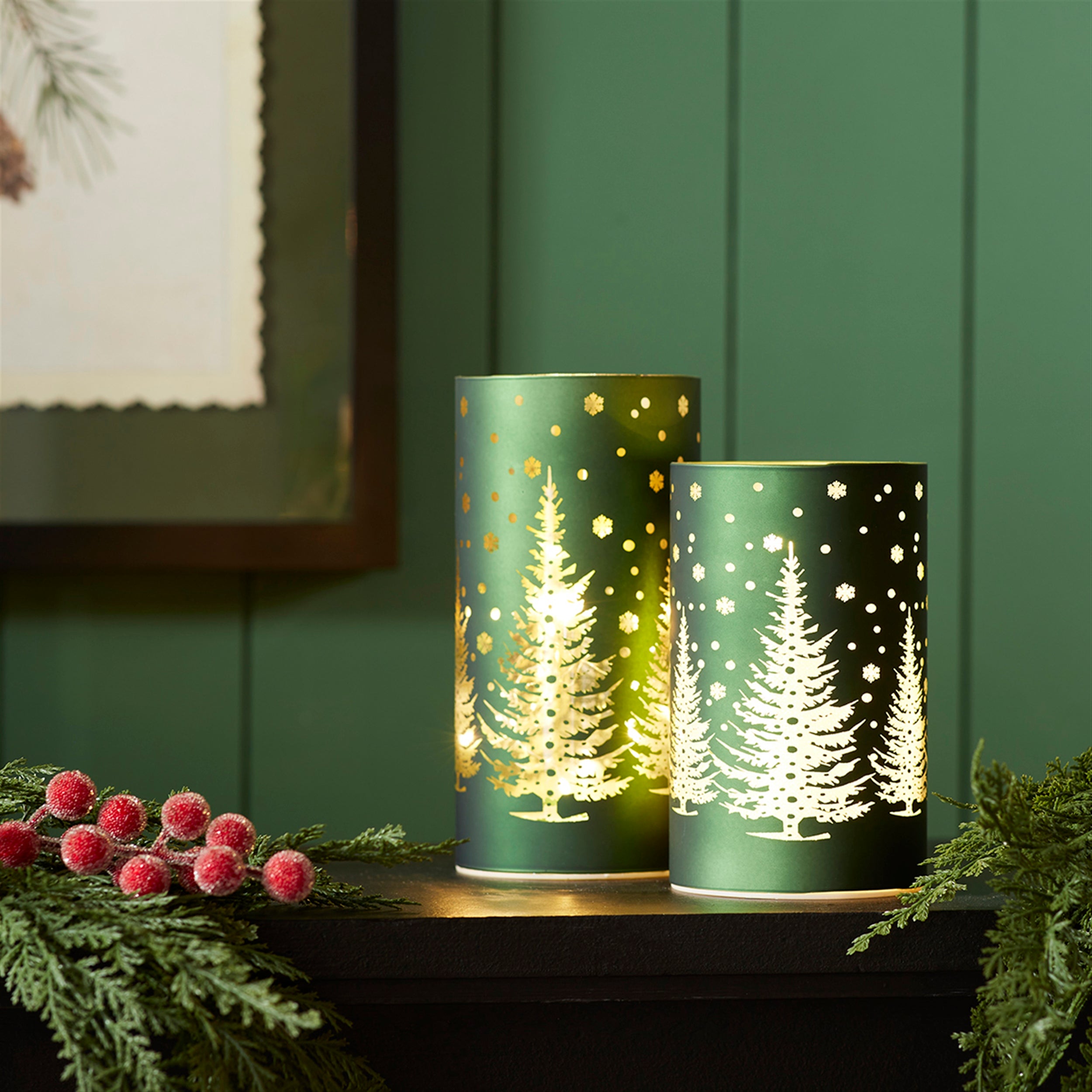 Two green tree luminaries glowing on a dark mantel with pine and red berries
