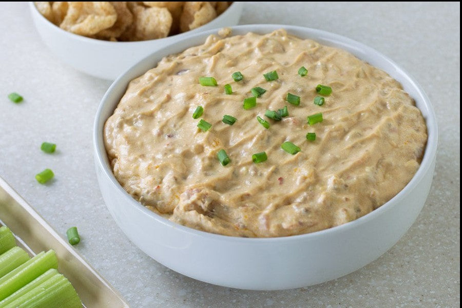 Bowl of Hatch Chile Cream Cheese Dip topped with green onions, with pork rinds and celery