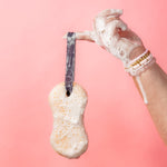 Hand holding the Sweet Grace Body Wash Sponge by its ribbon strap against a pink background