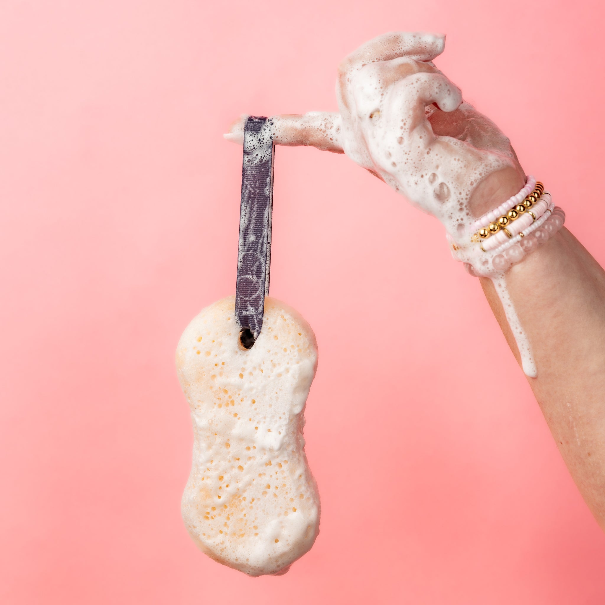 Hand holding the Sweet Grace Body Wash Sponge by its ribbon strap against a pink background
