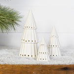 Three white ceramic light-up Christmas trees with punched-hole patterns grouped on a snowy wood shelf