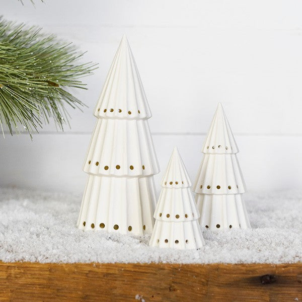 Three white ceramic light-up Christmas trees with punched-hole patterns grouped on a snowy wood shelf