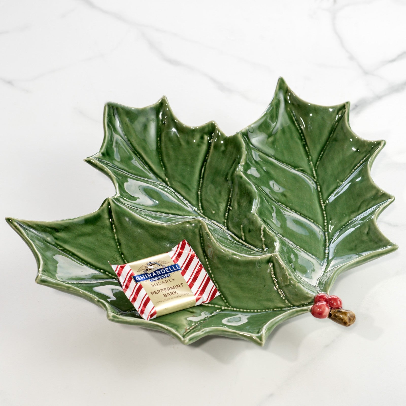 Green ceramic holly leaf plate with red berry accents holding a wrapped chocolate on marble