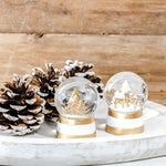Two mini snow globes with gold deer and tree scenes on white and gold bases beside frosted pinecones