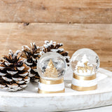 Two mini snow globes with gold deer and tree scenes on white and gold bases beside frosted pinecones