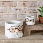 Two white Santa face planters with black wire glasses and rosy cheeks, large and small, on a wood shelf