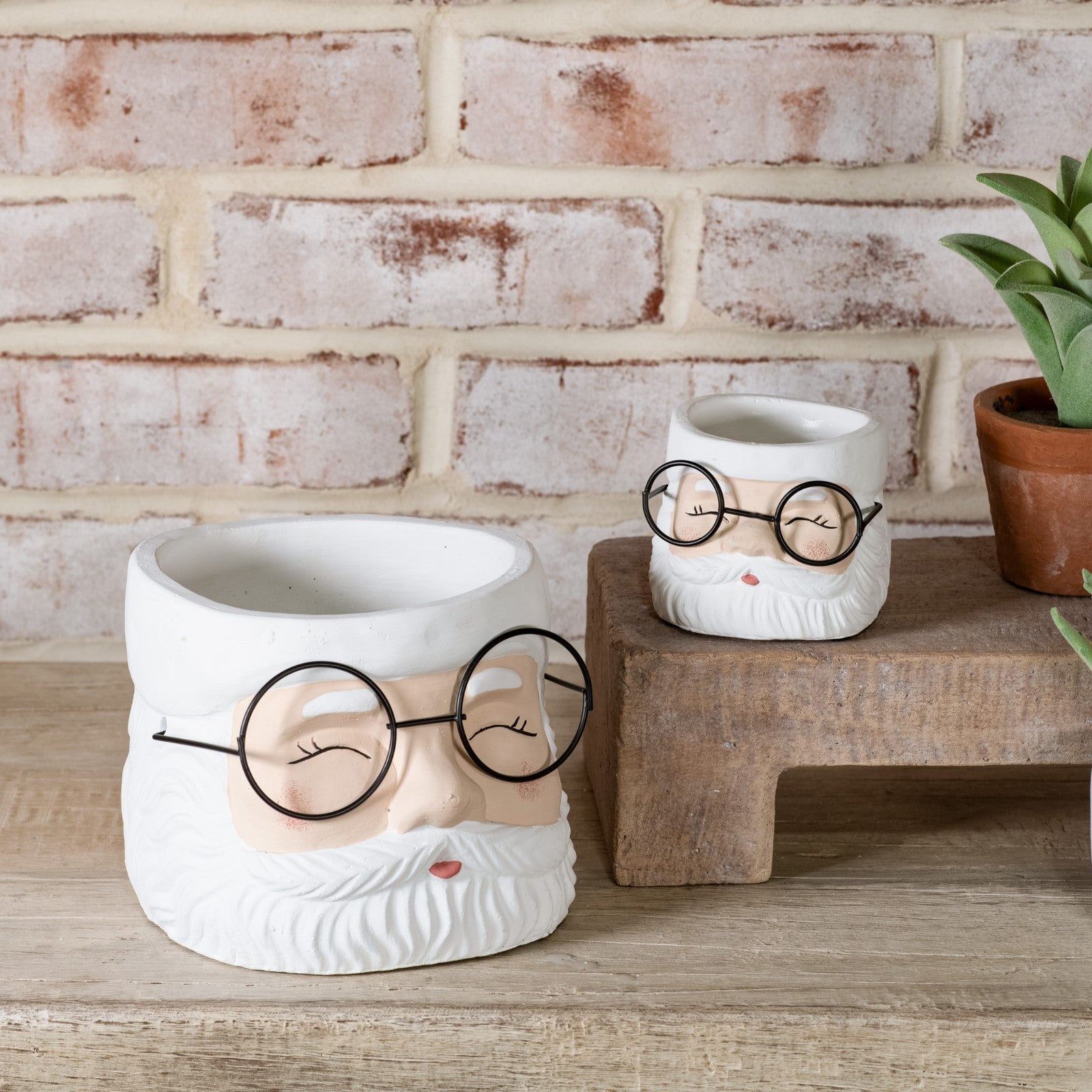 Two white Santa face planters with black wire glasses and rosy cheeks, large and small, on a wood shelf