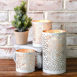 White metal lanterns with cut-out snowflake patterns glowing warm, grouped with a small potted pine