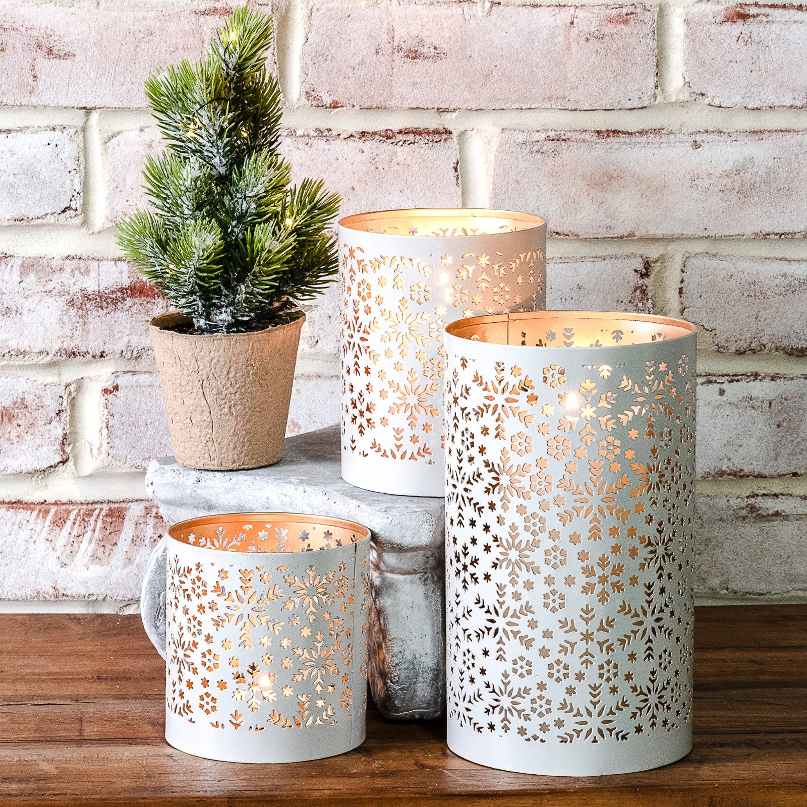 White metal lanterns with cut-out snowflake patterns glowing warm, grouped with a small potted pine