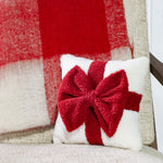 Small white textured pillow with a plush red bow and ribbon band, set against a red plaid throw