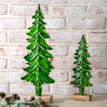 Two glossy green carved wood evergreen trees on natural wood bases, tall and short, against white brick