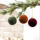 Velvet ball ornaments in forest green, rust orange and burgundy hanging from a frosted pine branch