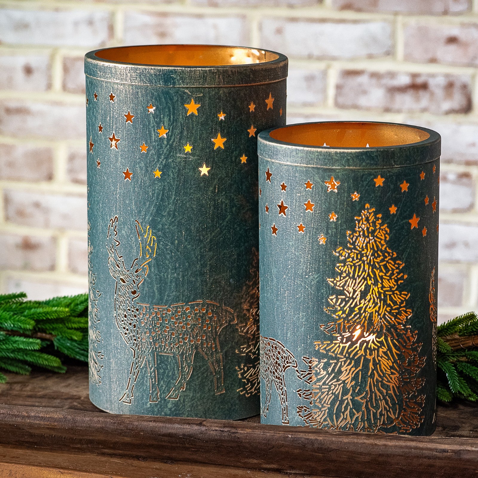 Two dark green lanterns with gold deer, forest and star cutouts glowing on a wood shelf