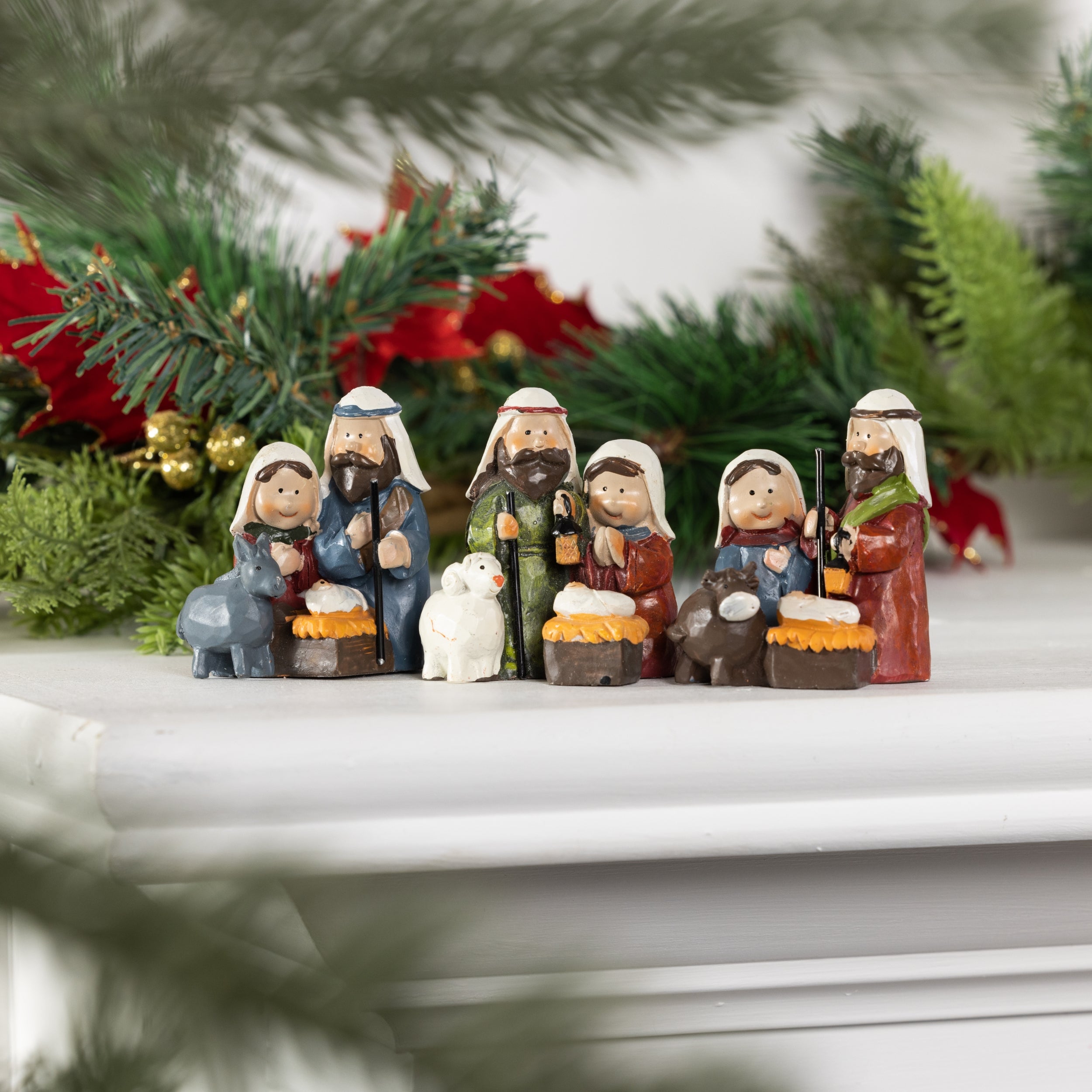 Mini nativity figurines lined up on a white mantel with pine and red poinsettia garland