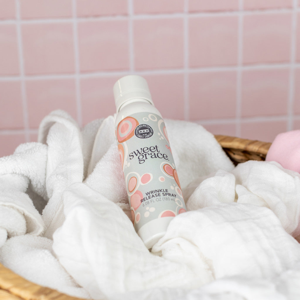 Sweet Grace Wrinkle Release Spray resting on white towels in a woven basket against a pink tiled wall