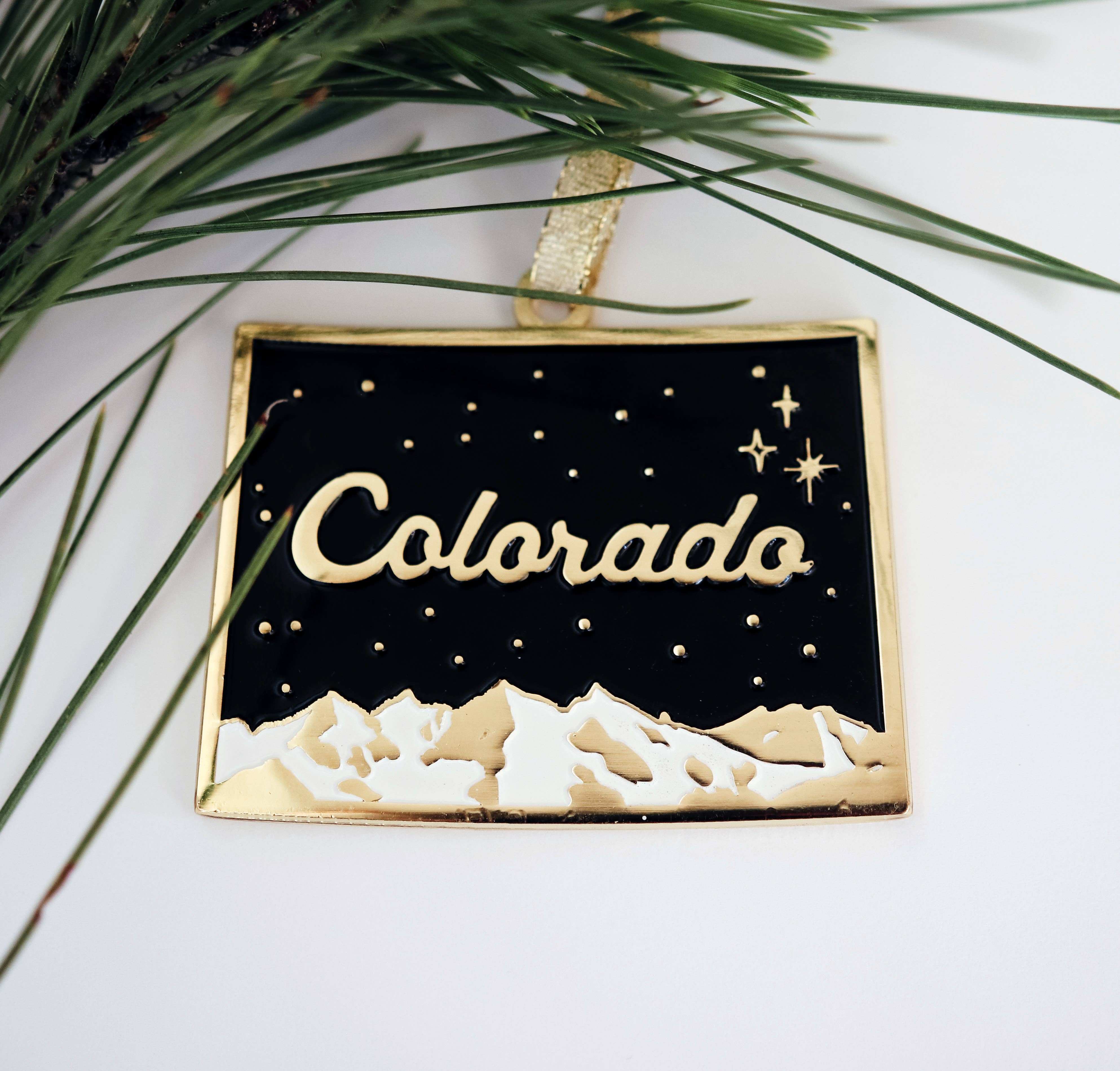 Colorado night sky enamel ornament with gold script lettering, shown beside pine needles