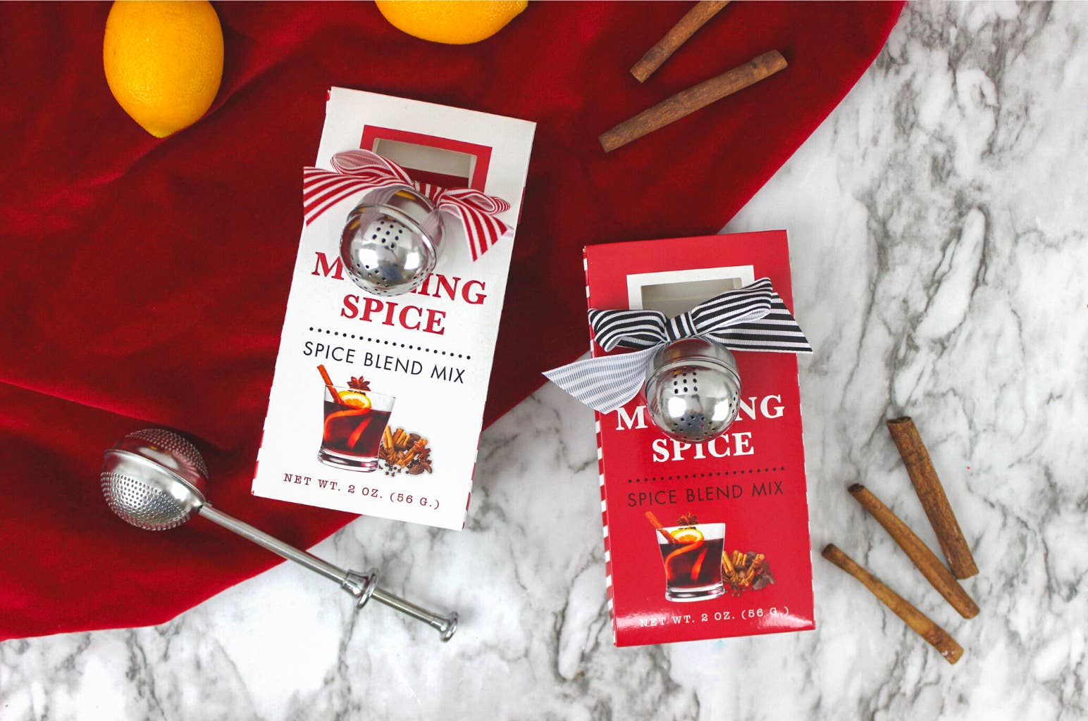 Two mulling spice boxes with striped ribbons and tea ball infusers on a red cloth with cinnamon sticks
